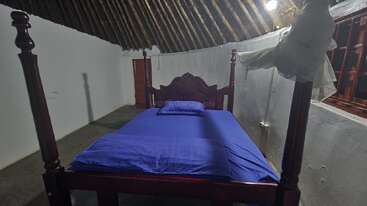 Uma cama de madeira de dossel com lençóis e travesseiros azuis fica em um quarto simples com paredes brancas, um teto inclinado de palha e uma janela.