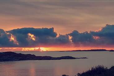 Eine ruhige Sonnenuntergangsszene über stillen Gewässern, mit dramatischen Wolken, die in goldenen und orangefarbenen Tönen leuchten. Felsinseln und Büsche, die sich als Silhouetten abzeichnen, runden die friedliche Küstenlandschaft ab.