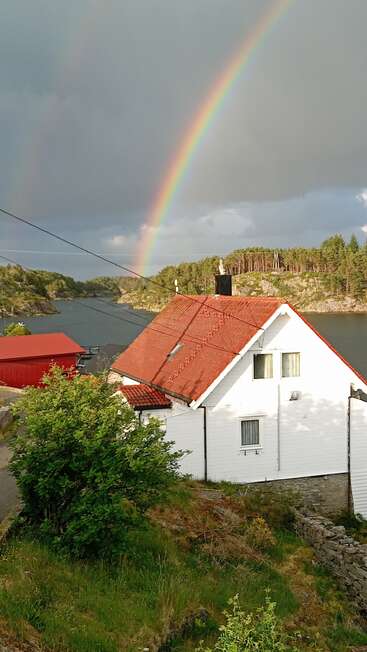 Ein leuchtender Regenbogen spannt sich über den bewölkten Himmel über einem ruhigen Fjord. Ein bezauberndes weißes Haus mit rotem Dach steht direkt am Wasser, umgeben von viel Grün.