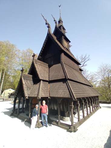 Zwei Menschen stehen vor einer hohen, kunstvoll verzierten Stabkirche aus Holz mit steilen Dächern und Drachenkopf-Schnitzereien, umgeben von Bäumen unter einem strahlend blauen Himmel.