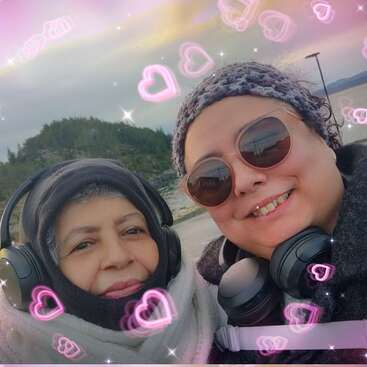 Two women bundled in winter clothing and headphones smile for a selfie outdoors. Pink heart graphics decorate the image, creating a warm, affectionate, and joyful atmosphere.