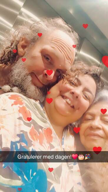 Three smiling people pose closely together, surrounded by floating red heart graphics. The photo features cheerful birthday wishes in Norwegian: “Gratulerer med dagen!❤️🎂🎉” taken inside an elevator.