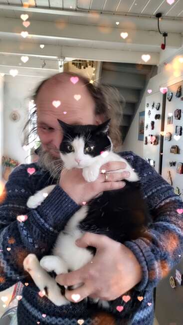A man lovingly holds a black and white cat in a cozy home. Heart-shaped filters float around, creating a warm, affectionate, and joyful atmosphere.