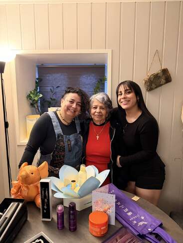 Three women smile together indoors, standing behind a table with beauty products, a stuffed orange animal, a gift card, and purple sashes. It appears celebratory.