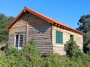 A rustic wooden cabin with a red sloped roof stands surrounded by green shrubs. The house has green shutters and white framed doors, under a clear blue sky.