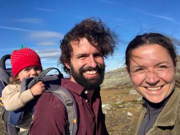 Eine glückliche Familie in der Natur: Ein bärtiger Mann trägt ein Kind in einer Rückentrage, begleitet von einer lächelnden Frau. Strahlend blauer Himmel und Berge.