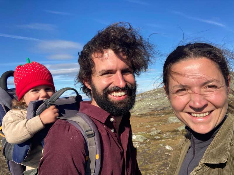Une famille heureuse en plein air : un homme barbu porte un enfant dans un sac à dos porte-bébé, accompagné d'une femme souriante. Un ciel bleu éclatant et des montagnes.