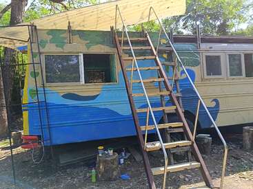 A school bus converted into a home, painted with blue, green, and yellow designs. A wooden staircase leads to a rooftop deck under a canopy.