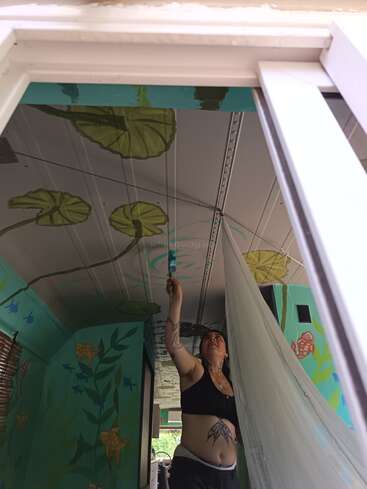 A woman paints green lily pads and aquatic plants on the ceiling of a room, surrounded by colorful murals and fish designs, creating an underwater theme.