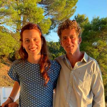 Ein fröhlicher Mann und eine fröhliche Frau stehen zusammen draußen und lächeln sich warm an. Die Sonne scheint auf sie, mit grünen Bäumen und blauem Himmel im Hintergrund, was eine friedliche, glückliche Szene schafft.