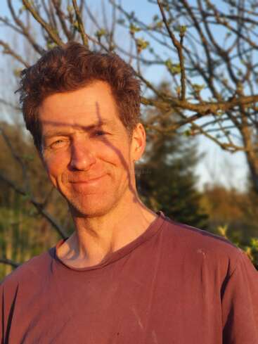 Un homme se tient debout à l'extérieur, souriant chaleureusement sous les rayons dorés du soleil. Les branches des arbres projettent des ombres ludiques sur son visage. L'arrière-plan est composé d'arbres en bourgeons et de couleurs douces et naturelles.