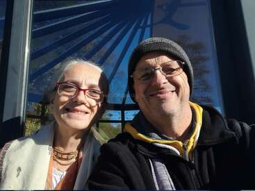 Una sonriente pareja de ancianos se toma un selfie al aire libre en un día soleado. Ambos llevan gafas y ropa de abrigo, con motivos geométricos azules que se reflejan en la ventana del fondo.