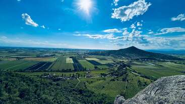 Uma vista deslumbrante da paisagem sob um sol forte, com campos verdes, terras cultivadas, uma montanha ao longe, nuvens espalhadas e um céu azul vibrante que se estende pelo horizonte.