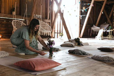 A woman sits on the floor arranging flowers in a cozy, sunlit barn with cushions, yoga mats, swings, and peaceful decorative lights creating a tranquil atmosphere.