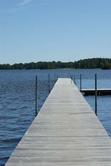A long wooden dock extends over calm blue water beneath a clear sky. Trees line the distant shore, creating a peaceful lakeside scene, perfect for relaxing.