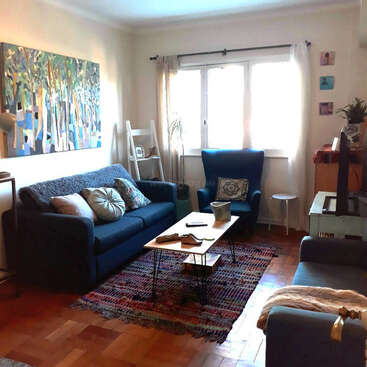 Das Bild zeigt ein gemütliches Wohnzimmer mit einer blauen Couch, einem Sessel, einem Couchtisch und einem Teppich, mit einem Fenster, Kunstwerken und einem Fernseher, die eine warme und einladende Atmosphäre ausstrahlen.