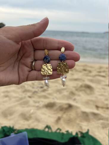 Eine Hand hält ein Paar elegante Ohrringe mit blauen Steinen, strukturierten Goldelementen und Perlentropfen. Im Hintergrund sieht man einen Sandstrand und das Meer.