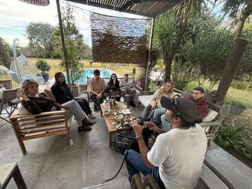 Eine Gruppe von sieben Leuten sitzt draußen auf einer überdachten Terrasse zusammen, genießt Essen und Getränke, während im Hintergrund ein Garten und ein Swimmingpool zu sehen sind.
