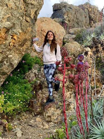 A woman stands on rocky terrain, smiling, holding a phone. She's surrounded by succulents and red flowering plants, dressed in athletic wear, enjoying a nature hike.