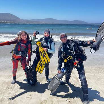 Three scuba divers stand smiling on a sandy beach, holding fins and gear, with the ocean and distant mountains behind them under a bright blue sky.