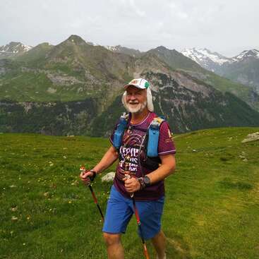 Un homme âgé et joyeux fait une randonnée en montagne, équipé de bâtons de trekking, d'une casquette, de matériel d'hydratation et de vêtements colorés. Des sommets enneigés et des collines verdoyantes l'entourent.