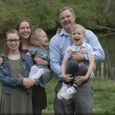 A happy family of five poses outdoors by a pond, smiling and laughing together. Lush green trees and calm water create a peaceful, joyful background scene.