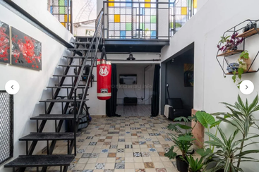 Dieses Bild zeigt ein offenes, modernes Loft mit einer Metalltreppe, einem hängenden Boxsack, einem bunt gefliesten Boden, Zimmerpflanzen, Wandkunst, Regalen und einem Buntglasbalkon.