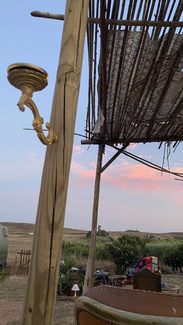 A rustic outdoor seating area with wooden poles, a patterned canopy, countryside view, cozy furniture, plants, and a golden wall sconce at sunset under a pastel sky.