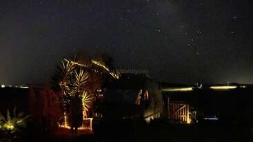 A cozy campsite illuminated by warm lights, features a van, palm plants, and a wooden fence under a dark, starry night sky with distant glowing horizon.