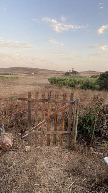 A rustic wooden gate stands amid dry grass, opening to a vast, open field with distant hills, sparse greenery, and a serene, cloudy blue sky.
