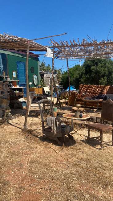 This image shows a rustic outdoor seating area with mismatched chairs, tables, and a couch under wooden pergolas, next to a green building surrounded by nature.