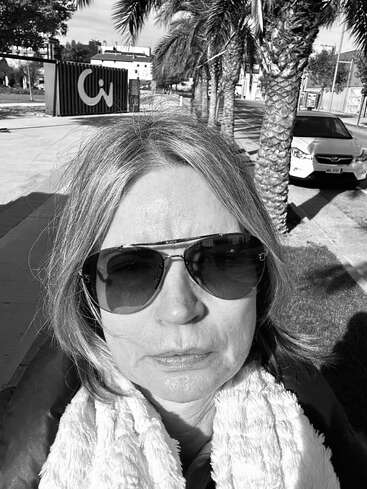 Uma mulher com cabelo liso e óculos escuros está ao ar livre, sob a luz do sol. Ela usa um cachecol fofo. Sombras e um carro são visíveis ao fundo. Foto em preto e branco.