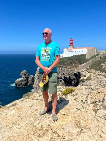 Un homme souriant portant des lunettes de soleil se tient sur une falaise rocheuse près de l'océan, en tenant son chapeau. Derrière lui se trouve un phare blanc avec un dôme rouge.