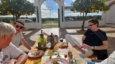 Trois hommes se détendent à une table en plein air après un repas, entourés de citronniers, de bouteilles de vin et de soleil, en profitant d'une conversation décontractée sur un patio lumineux.