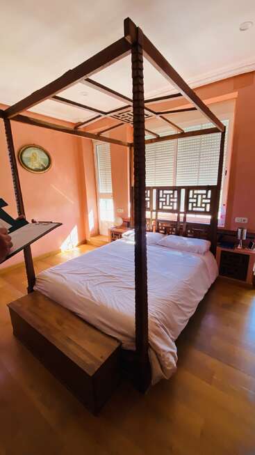 Une chambre à coucher confortable aux murs chauds de couleur pêche comprend un lit à baldaquin en bois, une literie blanche, une commode en bois, la lumière du soleil, des œuvres d'art au mur et des tables de chevet avec des lampes.