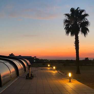 Un coucher de soleil serein avec un ciel coloré, la silhouette d'un palmier, l'enceinte d'une piscine, l'éclairage des allées et un paysage paisible et ouvert créent une atmosphère détendue et tranquille.