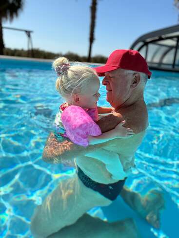 An elderly man in a red cap lovingly holds a young blonde girl with pink floaties in a bright blue swimming pool under a sunny sky.