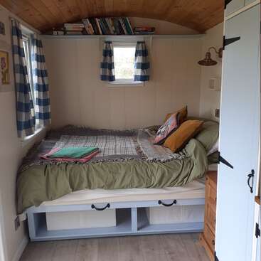 Cozy bedroom with a neatly made bed, green bedding, and pillows. Blue-checkered curtains, books on a shelf, under-bed storage, and wooden ceiling create a warm atmosphere.