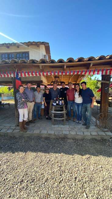 A imagem mostra um grupo de pessoas em frente a um prédio, com uma estrutura de madeira com uma bandeira do Chile e uma mesa com garrafas de vinho.
