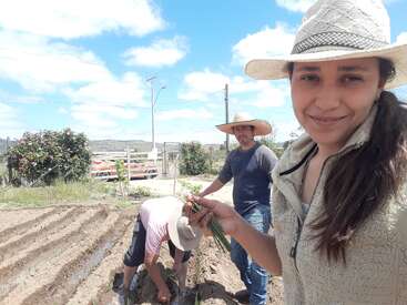 A imagem mostra uma mulher com um chapéu segurando cebolas verdes, em um jardim com duas pessoas trabalhando ao fundo em um dia ensolarado.