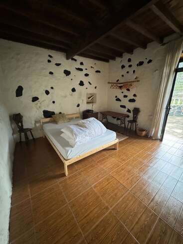 This cozy rustic bedroom features a wooden bed, simple furnishings, and a stone-accented wall. Large windows with curtains open to natural light and serene outdoor views.