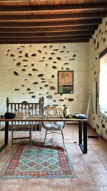 This image shows a rustic room with white stone walls, wooden beams, a cozy desk setup, rattan chair, colorful patterned rug, and a vintage travel poster.