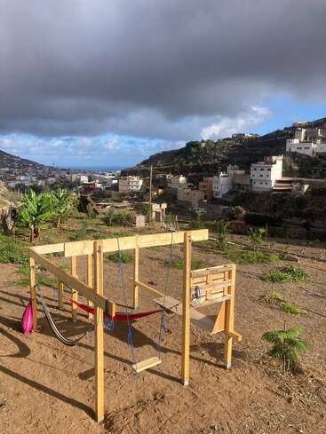 A handmade wooden playground structure sits on a dirt patch, surrounded by greenery. In the background, terraced hillside houses and dramatic clouds overlook a distant blue sea.