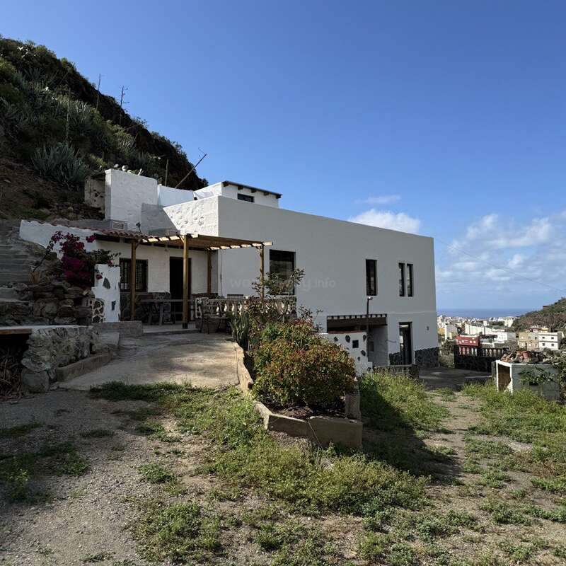 A modern white house with a patio sits on a hillside, surrounded by greenery. The sky is clear, overlooking a distant town and ocean views.