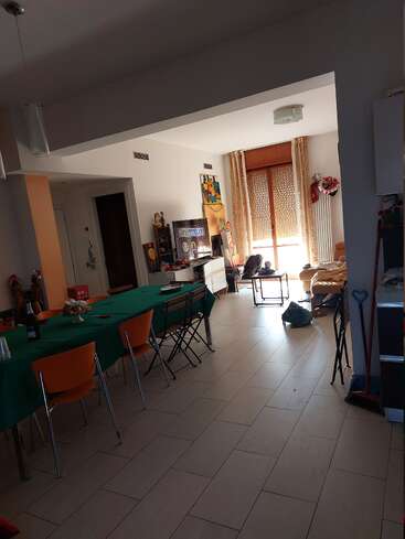 La imagen muestra una amplia habitación con una zona de comedor, con un mantel verde y sillas naranjas, junto a una sala de estar con un sofá y varios objetos.