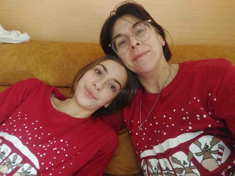 La imagen muestra a dos mujeres con camisas rojas a juego con dibujos blancos, posando para un selfie en un sofá marrón. La mujer de la izquierda tiene el pelo oscuro y la de la derecha, el pelo corto y oscuro.