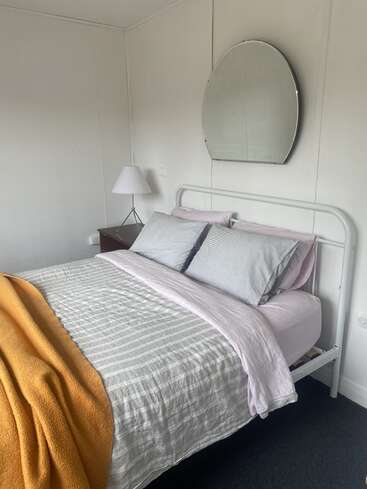 Ein ordentlich gemachtes Bett mit gestreifter Bettwäsche, hellrosa Laken und einer orangefarbenen Decke. Auf einem Beistelltisch steht eine weiße Lampe. Darüber hängt ein halbkreisförmiger Spiegel.