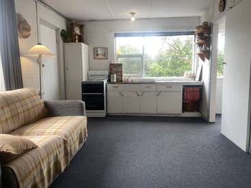 Ein gemütlicher, schlichter Wohnraum mit einem karierten Sofa, einer Stehlampe, einer Vintage-Küche, einem großen Fenster, weißen Schränken, offenen Regalen und viel Tageslicht, mit Blick auf die grüne Umgebung draußen.