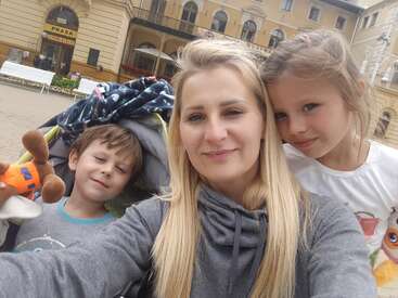 L'image montre une femme aux longs cheveux blonds posant pour un selfie avec deux enfants devant une rangée de bâtiments, probablement une rue de la ville.