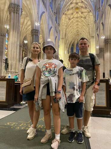 Une famille de quatre personnes se tient à l'intérieur d'une grande cathédrale aux hauts plafonds voûtés, aux colonnes ornées et aux vitraux, posant pour une photo lors de leur visite.